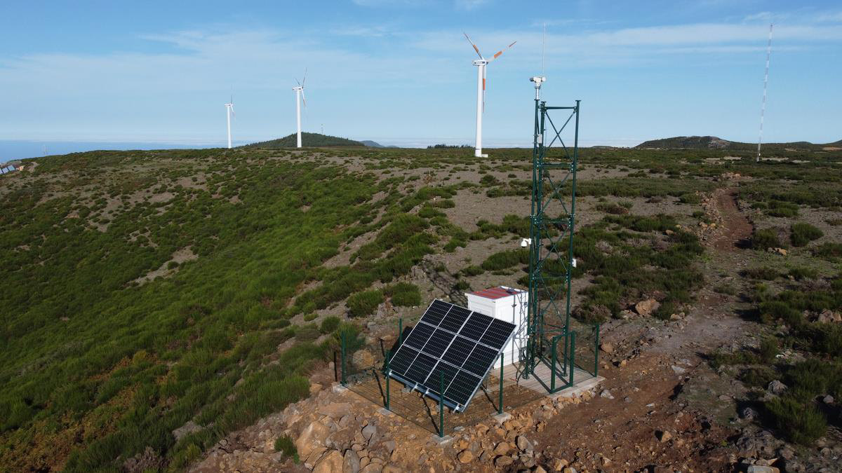 SafeForest tower with solar panels and equipment cabinet on a ridge in Madeira, wind turbines behind
