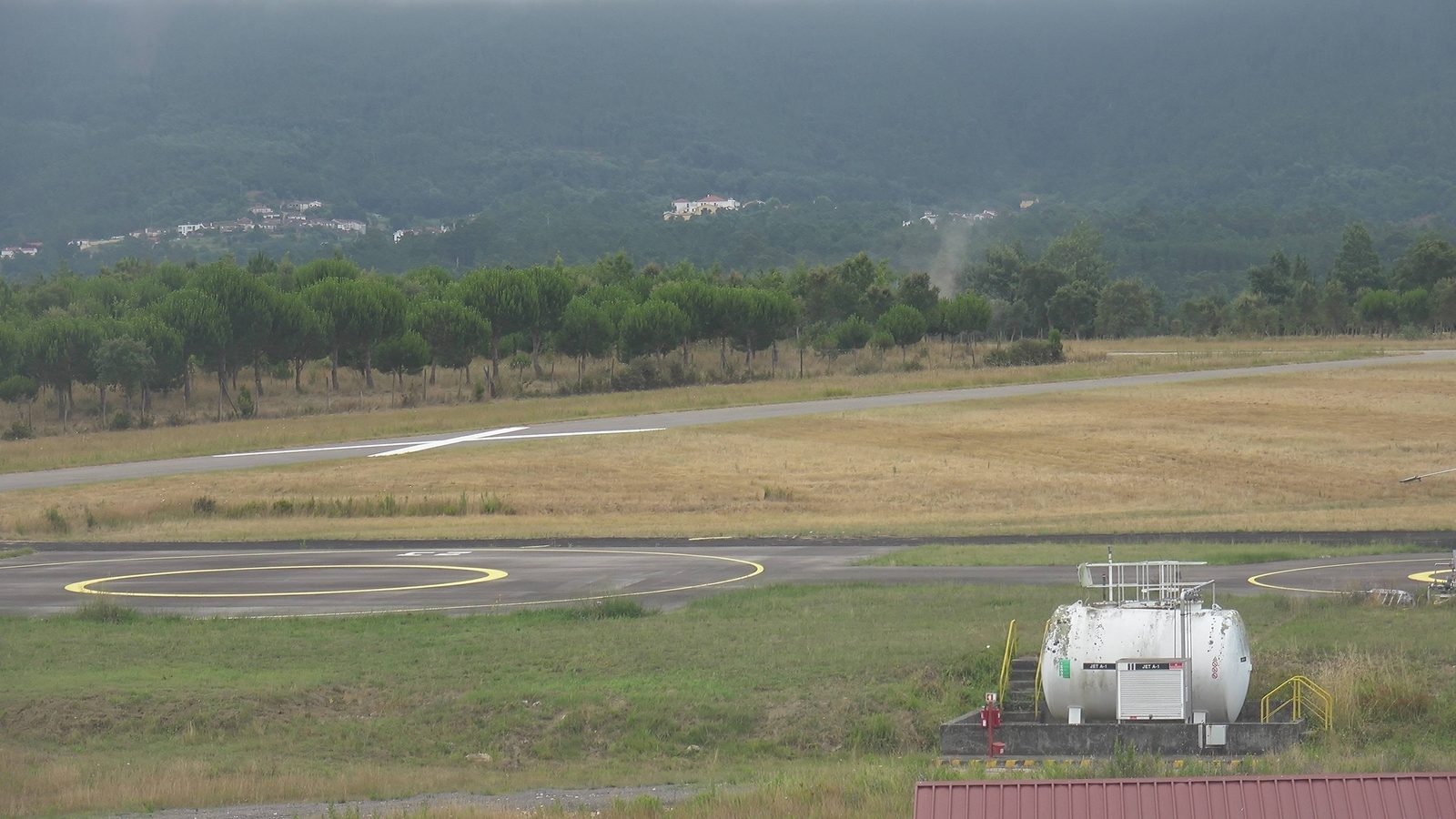 Camera frame: a thin smoke plume rising over the treeline beyond an airfield