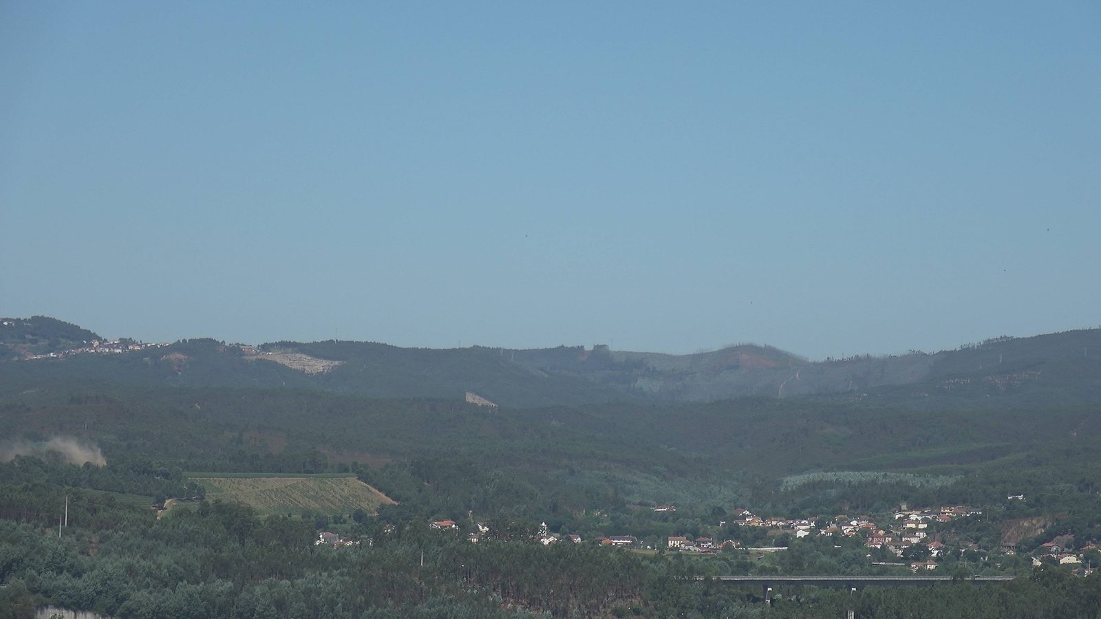 Camera frame: a faint smoke column rising from wooded hills above a river valley village
