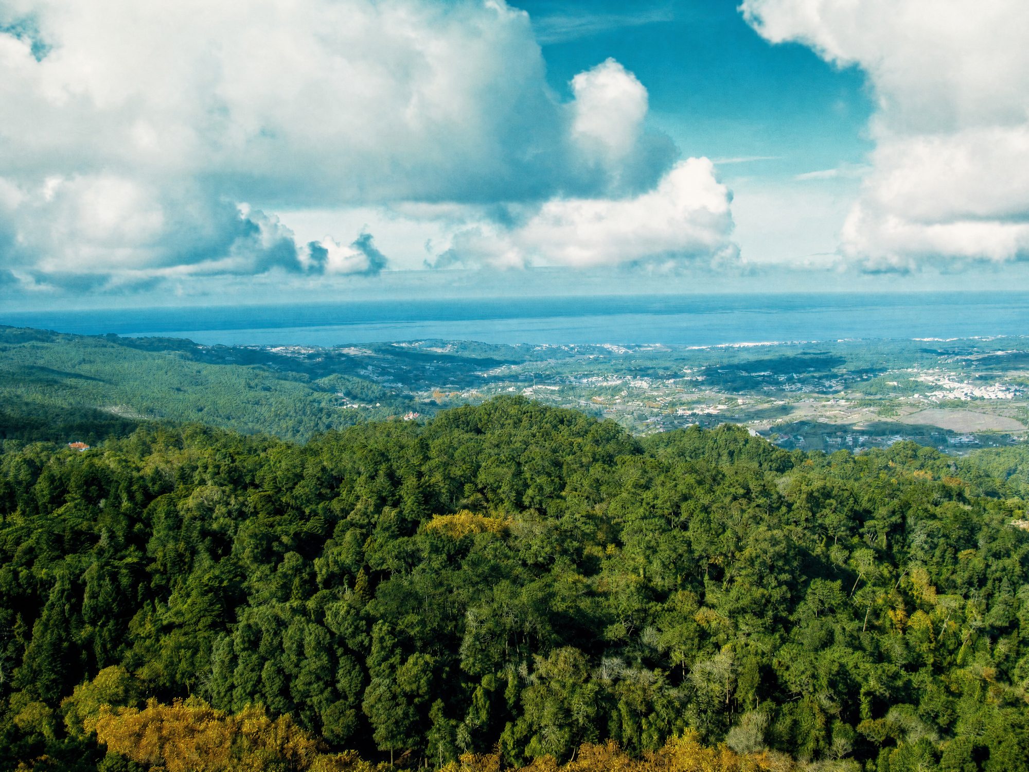 Aerial view over the forested Sintra mountains towards the Atlantic coast, under a sky of scattered clouds